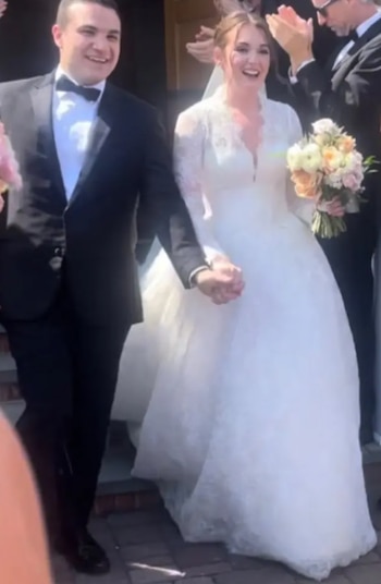 Un hombre con traje y pajarita camina de la mano de una mujer vestida de novia con un ramo de flores entre aplausos