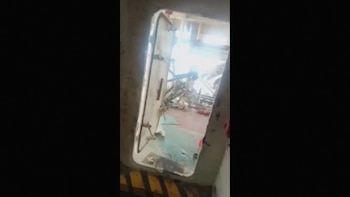 Puerta estanca metálica abierta con franjas amarillas y negras en el suelo y un espacio iluminado con maquinaria al fondo