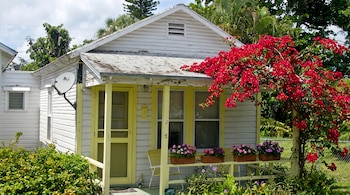 Casa pequeña de madera con revestimiento blanco, porche amarillo, techo de tejas, antena satelital, buganvilla roja y plantas en macetas.