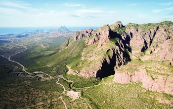 Vista aérea de montañas rocosas y vegetación árida, con caminos de tierra, y un área costera con mar y edificios en la lejanía