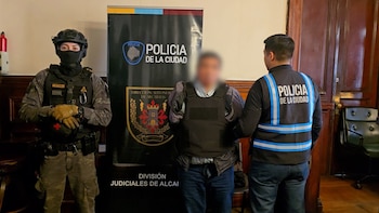 Un hombre con chaleco antibalas y rostro difuminado permanece custodiado por dos agentes de la Policía de la Ciudad frente a un cartel