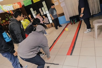 Un hombre inclinado lanza una pelota amarilla hacia varios pinos de plástico en una pista delimitada con cinta en el suelo