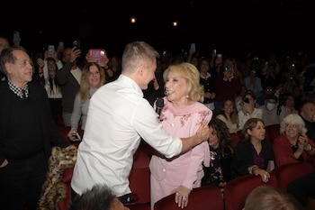 Una mujer rubia con traje rosa y un hombre en camisa blanca se sonríen y saludan en un teatro. El público sonriente los observa y graba con móviles