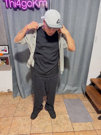 Thiago Medina, de pie, ajusta su camisa de rayas blanca y negra sobre una camiseta negra, gorra blanca y gafas, frente a cortinas grises y un letrero de neón