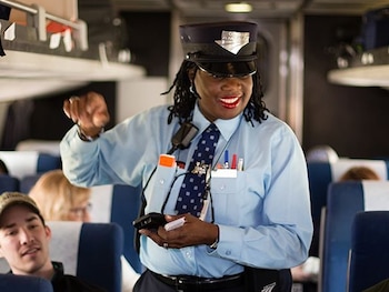 Una trabajadora afroamericana de Amtrak con uniforme azul y gorra sonríe mientras sostiene un dispositivo electrónico y tarjetas dentro de un vagón de tren