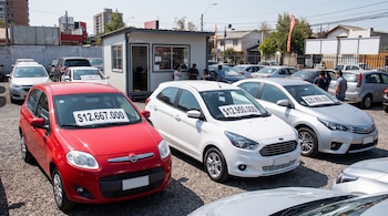 Tres automóviles de color rojo, blanco y gris presentan carteles de precios en sus parabrisas dentro de un predio de grava.