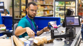 Hombre con gafas y delantal azul escaneando productos en una caja de supermercado. Se ven huevos, lácteos y otros artículos en el mostrador