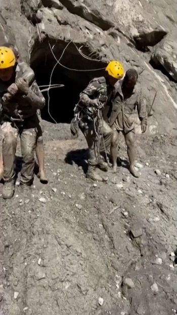 Rescatistas con cascos amarillos y uniformes cubiertos de barro ayudan a personas a salir de una abertura en la roca