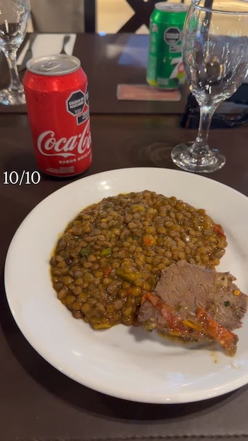 Plato blanco con estofado de lentejas y un trozo de carne, junto a latas de gaseosa Coca-Cola y una verde, y una copa vacía en una mesa oscura