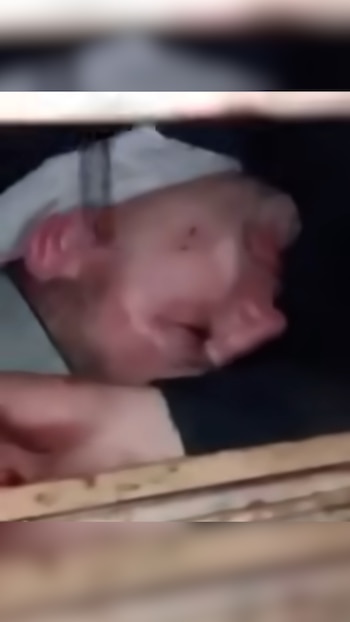 Rostro de un hombre con piel enrojecida, atrapado detrás de una estructura de madera horizontal. Parte de una prenda blanca rodea su cabeza. Fondo oscuro