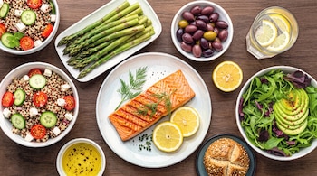 Ilustración en acuarela de un plato de salmón, ensaladas de quinua y aguacate, espárragos, aceitunas, aceite de oliva, pan y agua con limón.