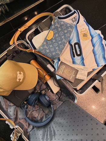 Maleta abierta con camiseta de Argentina número 10, pasaporte argentino, gorra beige Celine, auriculares azules, bolso Louis Vuitton y cargador blanco