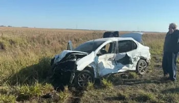 Automóvil sedán blanco con daños significativos en la parte delantera y lateral, sobre un campo de pasto seco bajo un cielo azul. Una persona de pie al costado