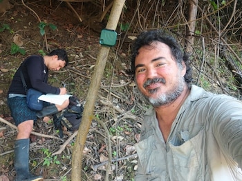 Dos hombres en un entorno natural. Uno con barba sonríe a cámara y el otro escribe en un cuaderno. Un dispositivo verde está sujeto a un árbol