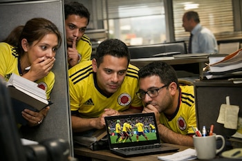 Cuatro personas con camisetas de Colombia observan un partido de fútbol en una laptop; uno hace un gesto de silencio y otra se cubre la boca.