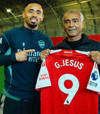 El futbolista Gabriel Jesus, con camiseta azul, sonríe y señala una camiseta roja del Arsenal con el número 9, sostenida por Romário, de cabello gris