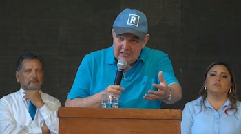 Un hombre con gorra azul con la letra R y camisa polo azul habla por un micrófono en un podio de madera, junto a un hombre y una mujer.