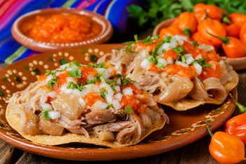 Dos tostadas con carne, cebolla, cilantro y salsa roja sobre un plato de barro, junto a chiles habaneros naranjas y una salsa.