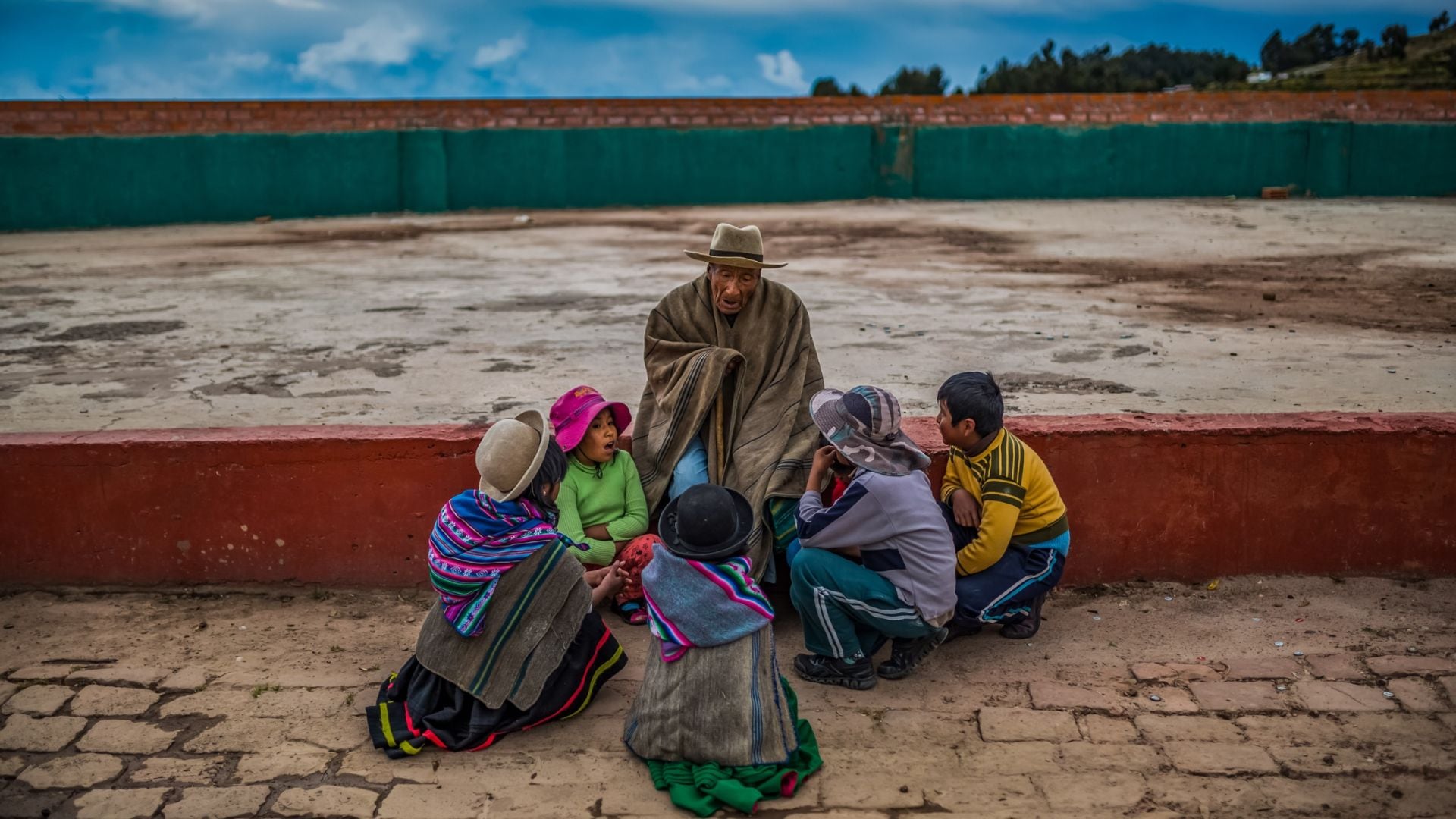 Un anciano andino comparte momentos de ocio con un grupo de niños, sentados en el suelo, transmitiendo posiblemente historias o conocimientos tradicionales. (MIDIS)