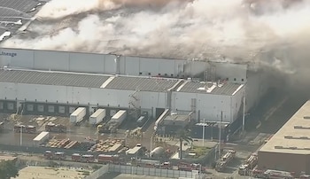 Vista aérea de un gran almacén con el nombre Lineage en su fachada, humo saliendo de su techo, camiones de bomberos y remolques de carga estacionados