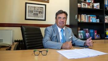 Retrato de Guillermo Marijuan, un hombre en traje gris y corbata azul, sentado en un escritorio de madera con gafas y papeles, fondo de estantería y oficina