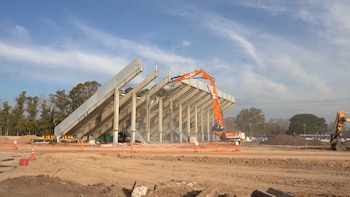 Una maquinaria de construcción naranja trabaja en la estructura de concreto de una tribuna en un terreno de tierra al aire libre