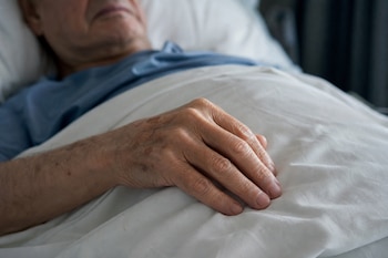 La mano de una persona mayor descansa sobre las sábanas blancas de una cama.