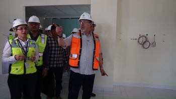 Un grupo de personas con cascos blancos de seguridad y chalecos reflectantes en una habitación. Un hombre señala hacia la derecha, se ven cables en la pared