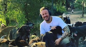 Un hombre sonriente con camiseta blanca y tatuajes acaricia a un perro negro mientras otros perros lo rodean en el campo
