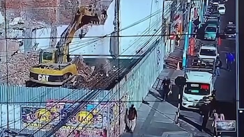 Vista aérea de una excavadora amarilla CAT en un sitio de demolición con escombros, junto a una valla metálica verde y una calle con tráfico y peatones