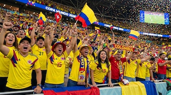 Multitud de aficionados colombianos con camisetas amarillas, banderas y bufandas animando en un estadio, con confeti cayendo y pantallas mostrando "Copa Mundo 2026".