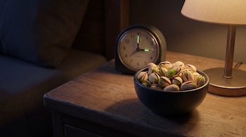 Un cuenco oscuro con pistachos, un reloj analógico y una lámpara encendida sobre una mesa de madera en una habitación oscura.