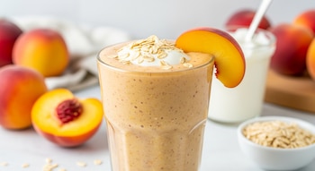 Un vaso de licuado de color naranja claro con avena y una rodaja de durazno. Al fondo, duraznos enteros, medio durazno y un recipiente de yogur.
