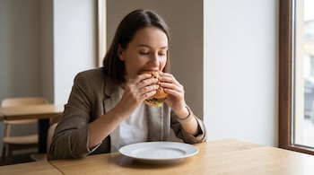 Mujer con chaqueta color café y camiseta blanca come una hamburguesa con lechuga y carne, sosteniéndola con ambas manos, en una mesa de madera clara.