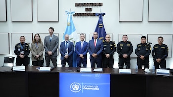 Un grupo de diez personas, incluyendo civiles en traje y personal policial en uniforme, posa de pie frente a banderas de Guatemala y un logo del Ministerio de Gobernación