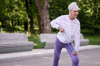 Mujer rubia con ropa deportiva morada y visera transparente, sujeta una banda de resistencia azul mientras se inclina en un parque con árboles y bancos