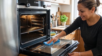Una mujer pasa un paño azul sobre la puerta de vidrio de un horno abierto en una cocina.