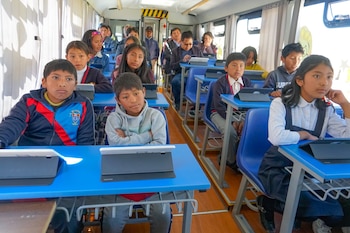 Grupo de niños y adolescentes sentados en escritorios azules con tabletas dentro de un autobús, con algunos adultos en la parte trasera
