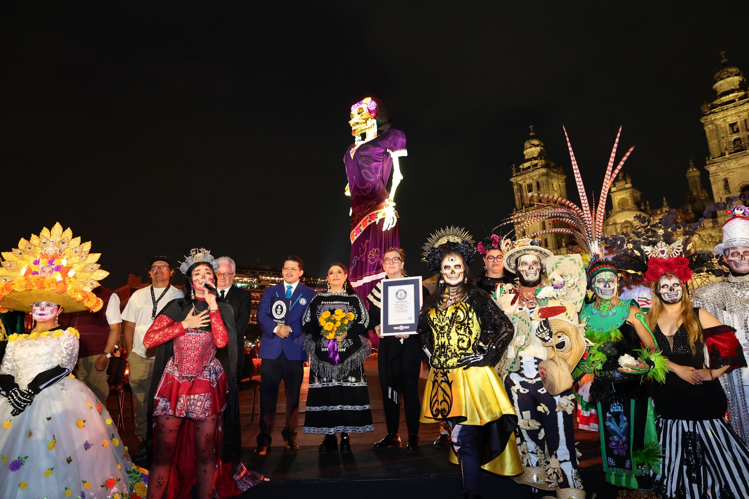 Autoridades de la CDMX y participantes caracterizados presentan un reconocimiento frente a una catrina monumental durante la inauguración de la ofrenda en el Zócalo de la Ciudad de México. (Gobierno CDMX)