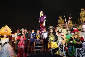 Personas caracterizadas de catrinas y funcionarios con un certificado posan de noche frente a una escultura monumental y una catedral iluminada