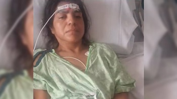 Una mujer recostada en una cama de hospital lleva una bata verde y un sensor en la frente