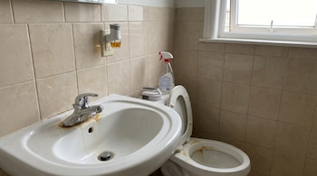 Baño con azulejos beige, lavabo blanco con grifo cromado y sarro, inodoro con tapa levantada y manchas de sarro, ambientador en enchufe y ventana.