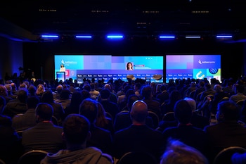 Multitud de personas de espaldas sentadas en un auditorio. Escenario con grandes pantallas azules que muestran logotipos de AmCham Argentina, texto y una mujer