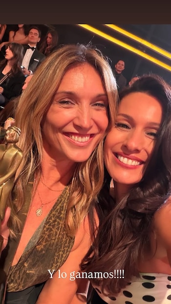 Primer plano de dos mujeres sonriendo; una con cabello rubio claro sostiene un trofeo dorado, la otra con cabello oscuro y un top de lunares posa a su lado