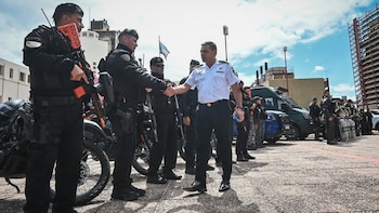 Un hombre con camisa blanca de policía estrecha la mano de un agente en uniforme táctico junto a motocicletas y vehículos policiales