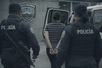 Dos policías uniformados escoltan a una mujer esposada. Un vehículo policial con la puerta abierta y el escudo de la Policía Nacional de Honduras son visibles.