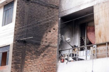 Pared de ladrillos ennegrecida, edificio con ventanas, balcón metálico con barandilla, objetos dañados y una cortina rosa en el interior