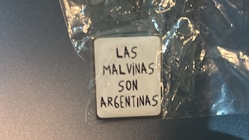 Pin cuadrado blanco con la leyenda 'Las Malvinas son Argentinas' dentro de una bolsa de plástico transparente sobre una superficie oscura