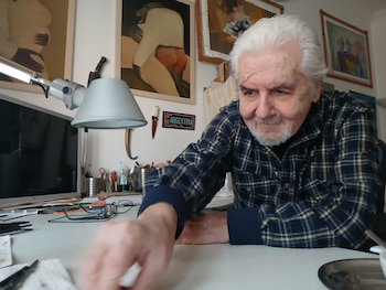 Hombre canoso de barba blanca con cicatriz sobre el ojo izquierdo, vistiendo camisa a cuadros, en su estudio con monitor, lámpara y obras de arte
