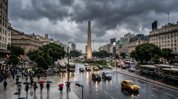 Avenida mojada con vehículos, peatones con paraguas en la acera, edificios urbanos, el Obelisco de Buenos Aires y nubes oscuras en el cielo.